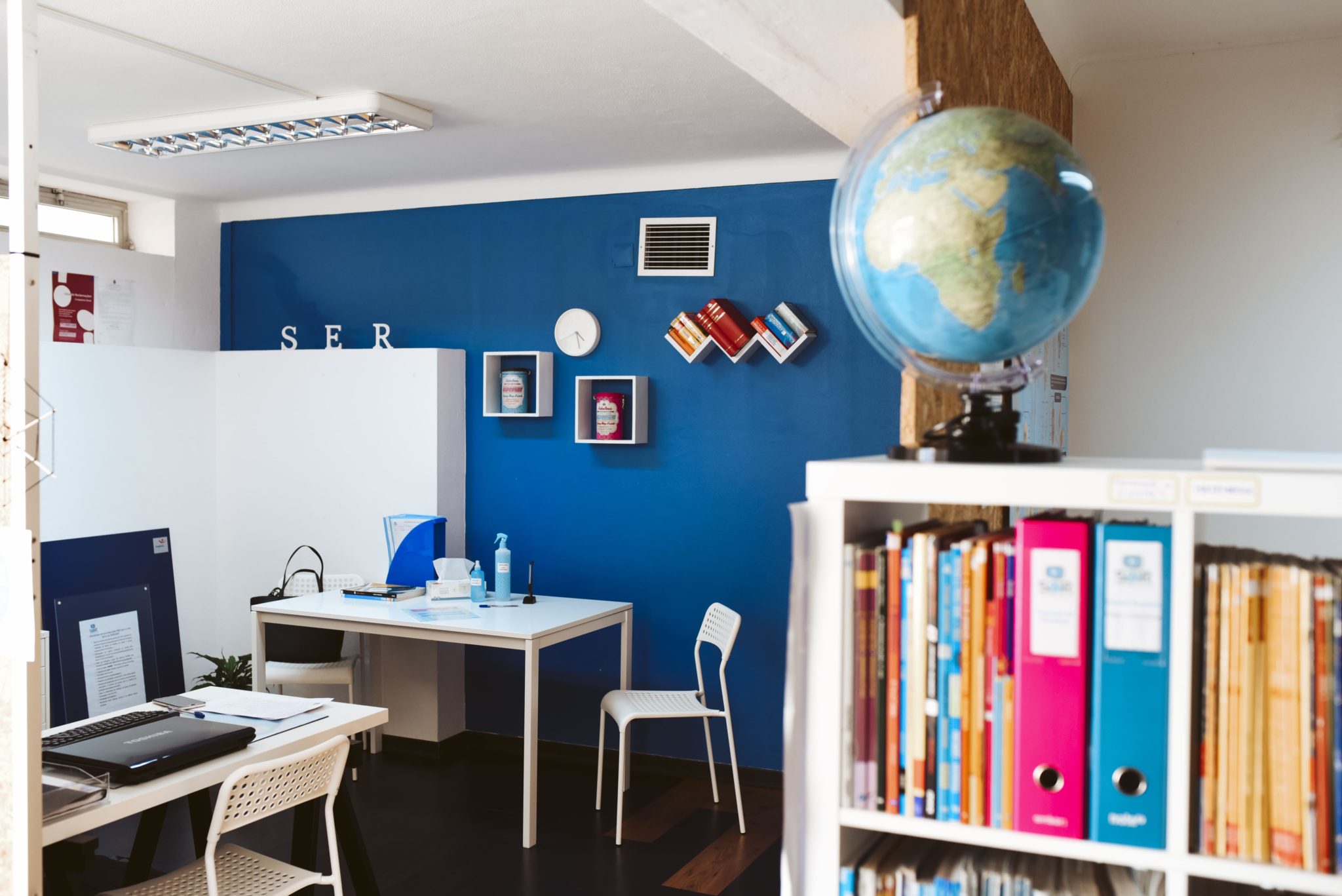 Sala de aula com livros, globos, mesas, cadeiras e computadores